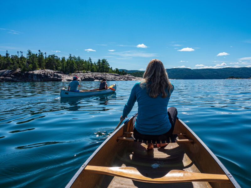 Lake Superior Paddle A woman paddles in the bow of a canoe with another canoe ahead on unusually calm waters in Lake Superior Provincial Park.