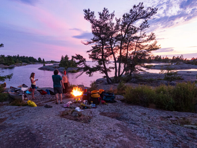 Fingerboard Islands Campfire A Canoe trip group stands around a fire on the shores of Georgian Bay as the setting sun sets the sky alight with purple colour.